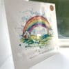 A print of a rainbow arching between two houses, named Under the Same Rainbow by Kathryn Callaghan. The 30cm print is presented flat in a clear cello bag with a golden KC sticker in the top right.