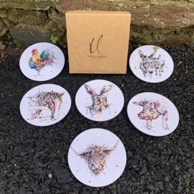 Six coasters featuring prints of farmyard animals by Kathryn Callaghan sit on a stone floor outside around a branded box.