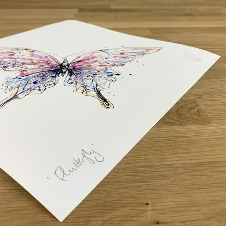 Photograph of Flutterfly, a print of a pink and blue butterfly by Kathryn Callaghan, which shows the print's title handwritten in the bottom left corner and the artist's signature in the bottom right, with space between to add a custom personalisation.