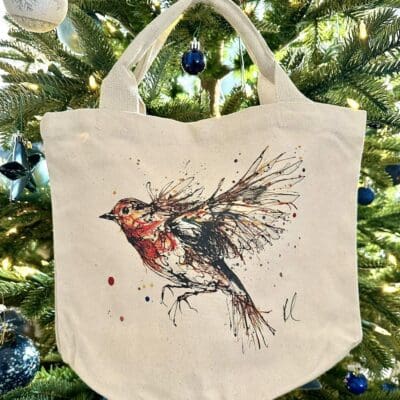 A canvas bucket tote bag featuring a painting of a robin in flight named Airborne by Kathryn Callaghan. The bag is hung from a Christmas tree.