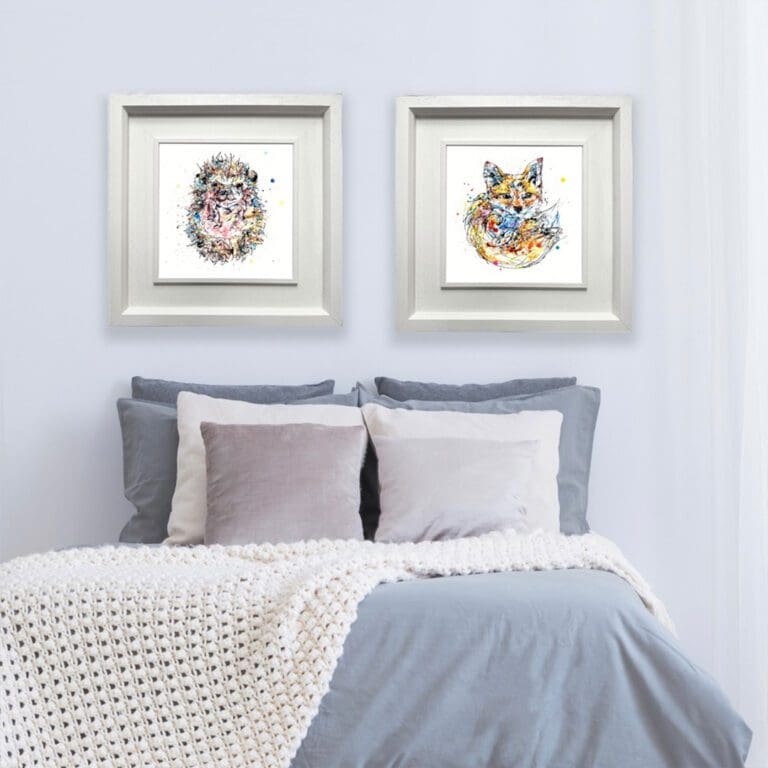 Two square prints are shown in Deluxe White Frames on a pale blue wall above blue and white bed cushions. The prints featured a curled up hedgehog named Pickles and a fox cub named Milkshake.