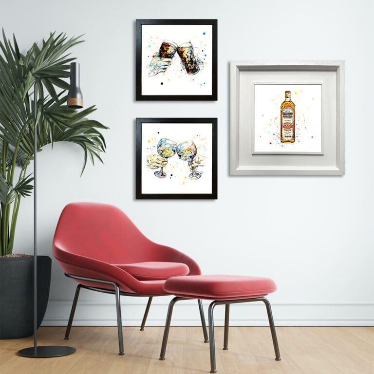 Three prints are shown in frames in a room with a pink chair and palm plant. The prints are Cheers which shows cheers-ing pinks in a standard black frame, Good Times which shows cheers-ing gin glasses in a Standard Black Frame, and Bushmills Irish Whiskey which shows a bottle of whiskey in a Deluxe White Frame.