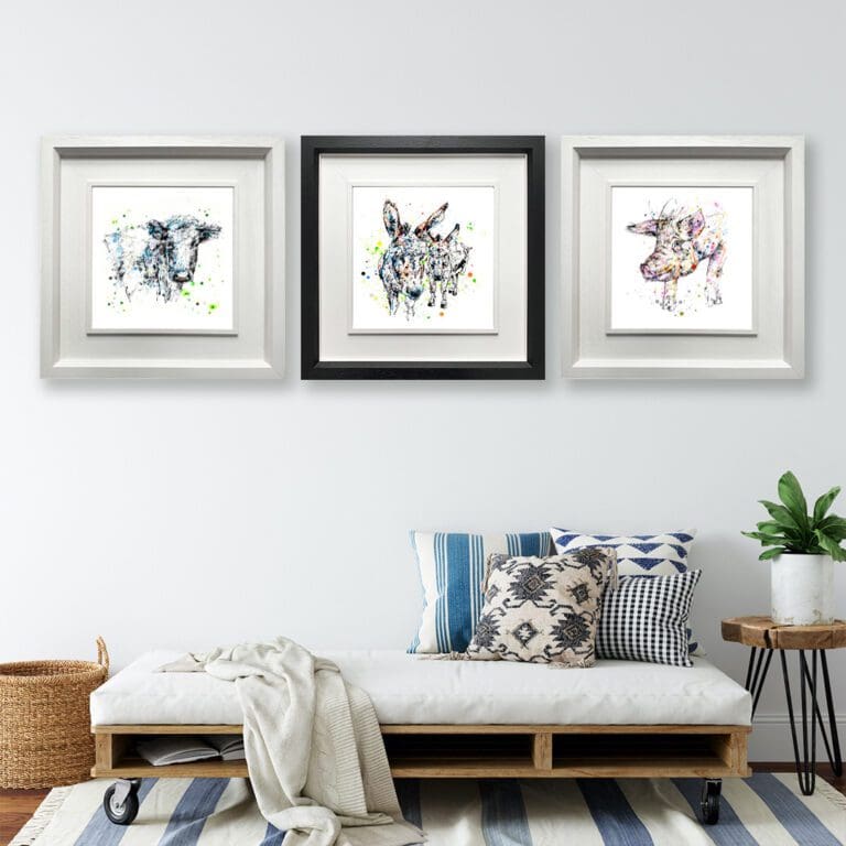 Three prints shown framed in situ on a white wall above a futon. The prints are Pastures New, which depicts a dairy cow in a deluxe white frame, Companions, which depicts a pair of donkeys in a deluxe black frame, and Truffles, which depicts a pig in a deluxe white frame.