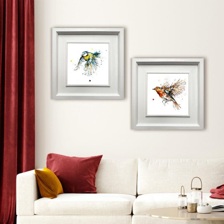 Two prints shown in Deluxe White Frames in a sitting room with cream walls and sofa and red velvet curtains. The prints are Airborne, which depicts a robin in flight, and Coat of Colour, which depicts a blue tit in flight. They are both flying left.