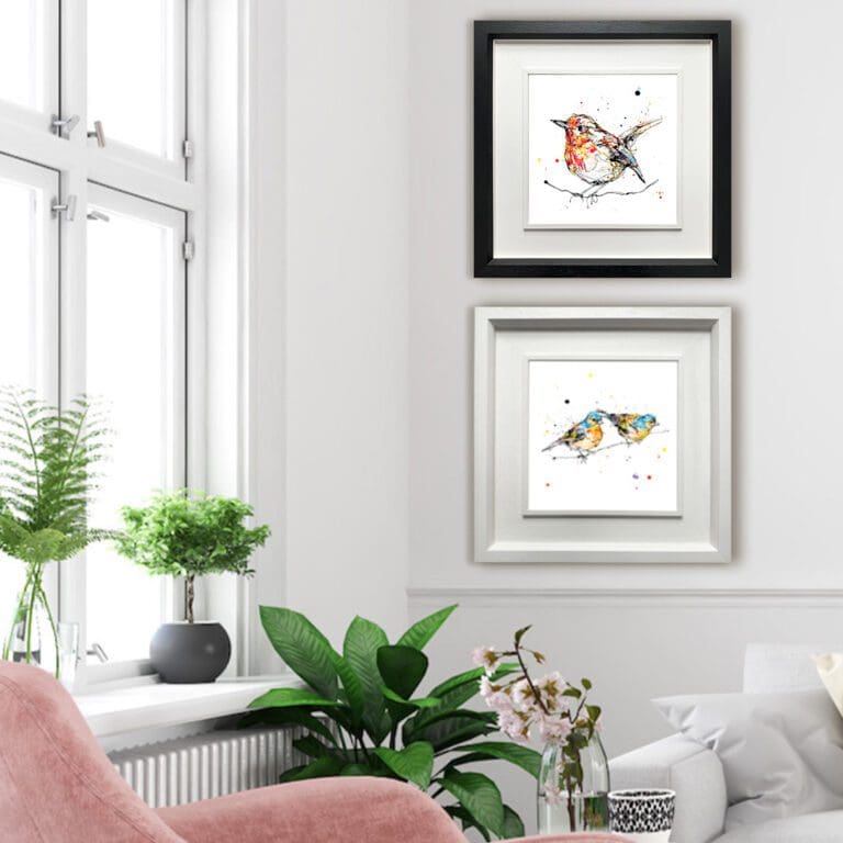 Two prints shown in Deluxe Frames in a white sitting room by a window with a pink chair and houseplants. The prints are Peekaboo, which shows a perched robin facing left in a deluxe black frame, and Did You See That? which depicts two chaffinches on a branch looking right in a deluxe white frame.