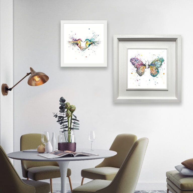 Two prints are shown in white frames in situ above a dining table with a plant. The prints are Forever, which depicts two hummingbirds flying in the shape of a heart, in a standard white frame, and Destiny, which depicts two butterflies facing one another, in a Deluxe White Frame.