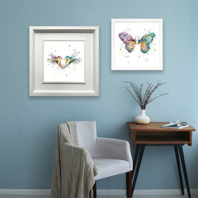 Two prints are shown in white frames in situ on a soft blue wall above a grey armchair and wooden side table. The prints are Forever, which depicts two hummingbirds flying in the shape of a heart, in a deluxe white frame, and Destiny, which depicts two butterflies facing one another, in a Standard White Frame.