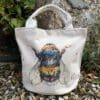A canvas bucket tote bag featuring a paining of a bee named Buzz by Kathryn Callaghan. The bag is sitting on gravel in front of a wall with plants growing from it.