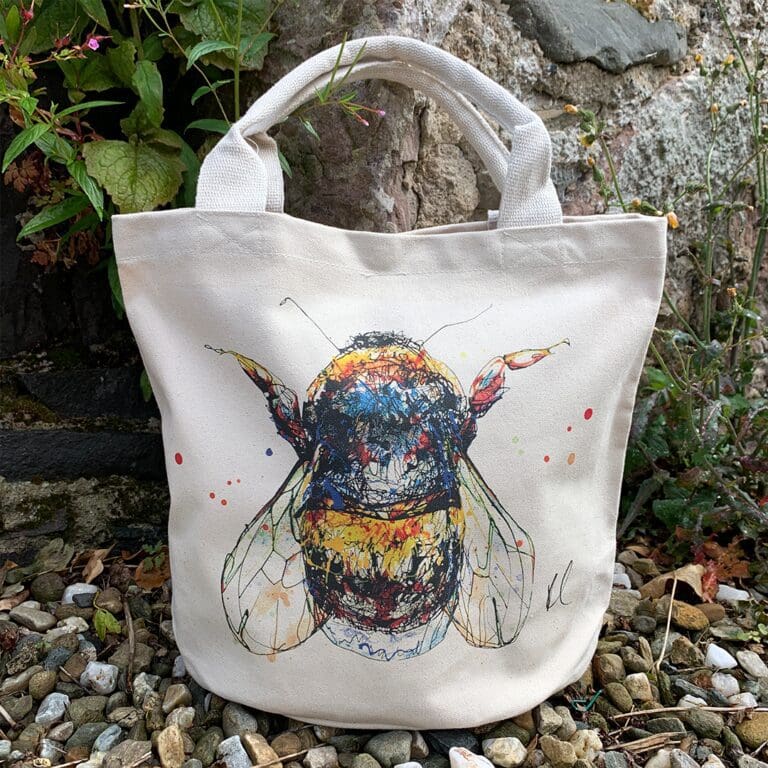 A canvas bucket tote bag featuring a paining of a bee named Buzz by Kathryn Callaghan. The bag is sitting on gravel in front of a wall with plants growing from it.