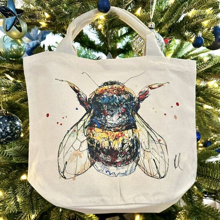 A canvas bucket tote bag featuring a painting of a bee named Buzz by Kathryn Callaghan. The bag is hanging from a Christmas tree.