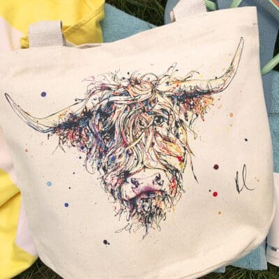 A canvas bucket tote bag featuring a paining of a highland cow named Harris by Kathryn Callaghan. The bag is sitting on blue and yellow beach towels on a grassy background.