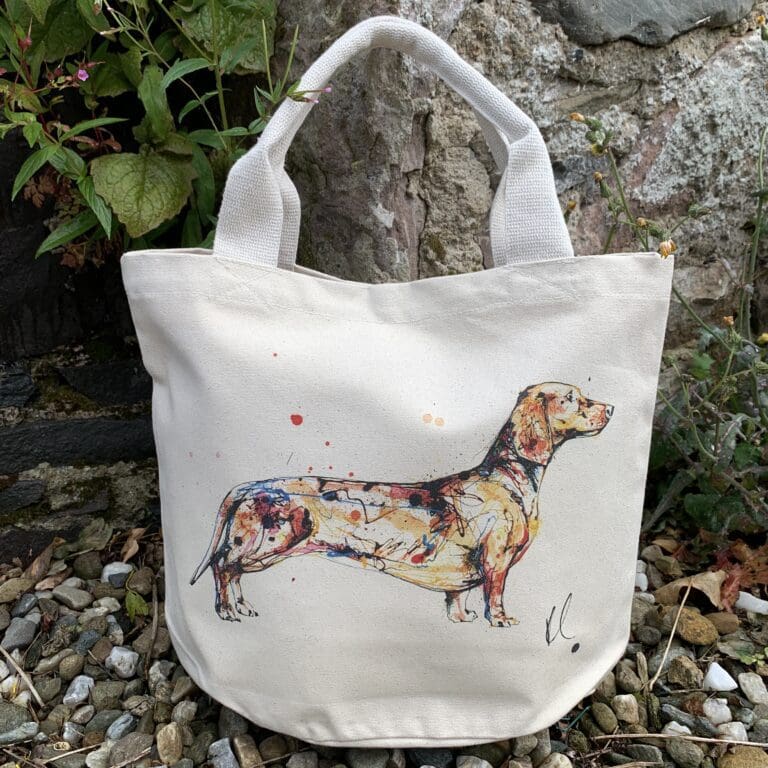 A canvas bucket tote bag featuring a paining of a dachshund named Hopeful by Kathryn Callaghan. The bag is sitting on gravel in front of a wall with plants growing from it.