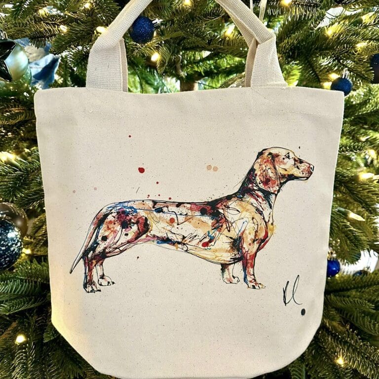 A canvas bucket tote bag featuring a paining of a dachshund named Hopeful by Kathryn Callaghan. The bag is hung from a Christmas tree.