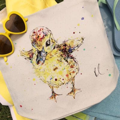 A canvas bucket tote bag featuring a paining of a duckling named Sherbet by Kathryn Callaghan. The bag is sitting on blue and yellow beach towels on a grassy background.