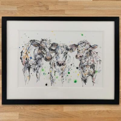 A Good Natter Original Cow Painting by Kathryn Callaghan, framed in a classic black frame with double white mount set on a wooden worktop.