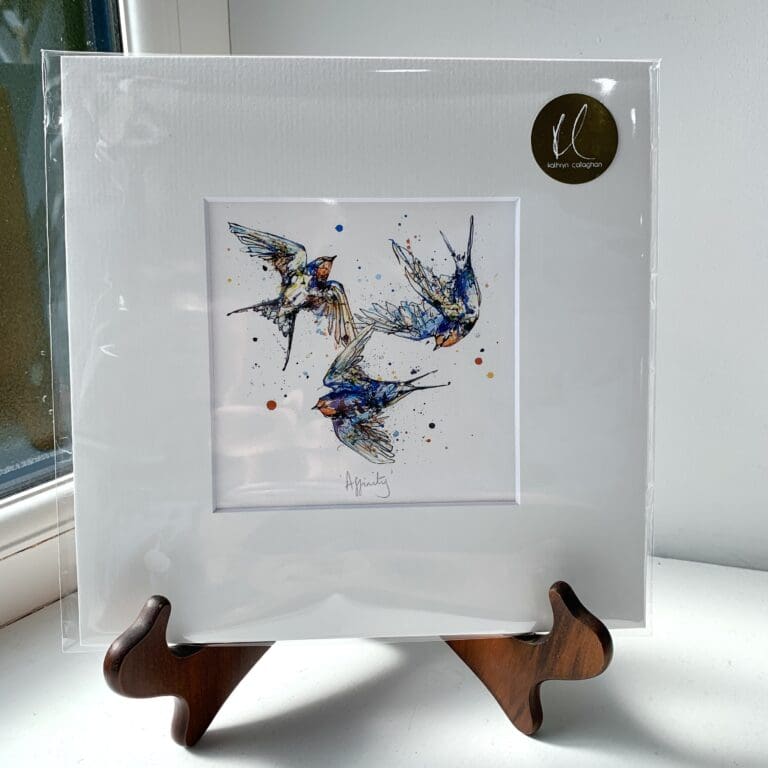 A miniature print of Affinity by Kathryn Callaghan, which features three swallows flying in a circle. The print is presented mounted at 23cm, in a clear cello bag with a gold KC sticker in the top right corner, and is displayed here on a small wooden easel in a windowsill.