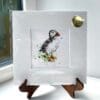 A miniature print of Atlantic Puffin by Kathryn Callaghan, which features a puffin standing tall in green grass. The print is presented mounted at 23cm, in a clear cello bag with a gold KC sticker in the top right corner, and is displayed here on a small wooden easel in a windowsill.