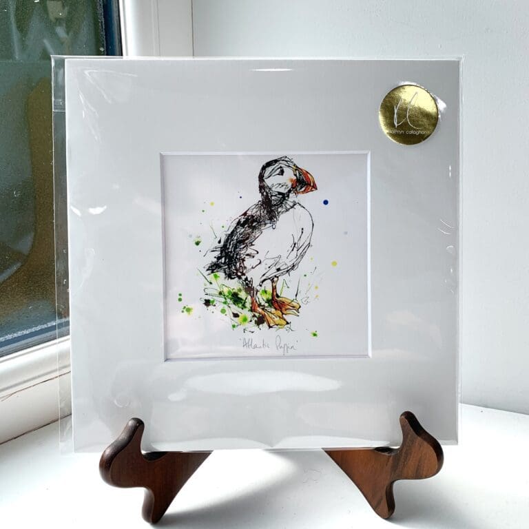 A miniature print of Atlantic Puffin by Kathryn Callaghan, which features a puffin standing tall in green grass. The print is presented mounted at 23cm, in a clear cello bag with a gold KC sticker in the top right corner, and is displayed here on a small wooden easel in a windowsill.