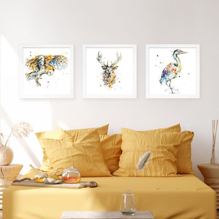 Three prints by Kathryn Callaghan shown displayed in standard white frames in situ on a white wall above a yellow bed. Sun shines in from the top left of the image. The prints are 'Hover', which features a hovering barn owl, 'Aura', which features a golden stag's head and torso, and 'Serenity', which features a colourful heron facing right.
