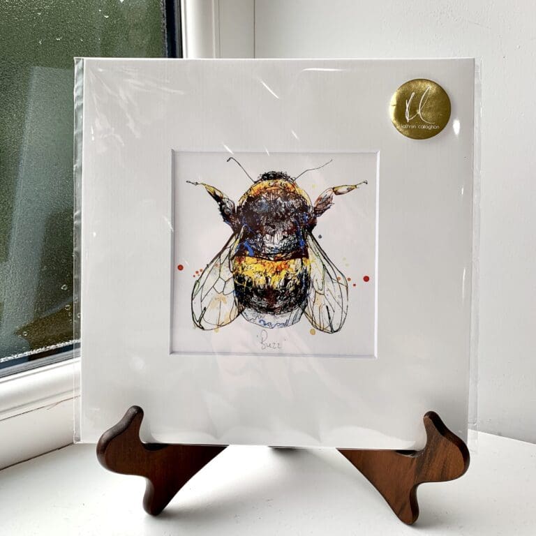 A miniature print of Buzz by Kathryn Callaghan, which features a bumblebee seen from above. The print is presented mounted at 23cm, in a clear cello bag with a gold KC sticker in the top right corner, and is displayed here on a small wooden easel in a windowsill.