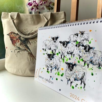 The Kathryn Callaghan 2024 Calendar, featuring a cover image of a painting of a flock of sheep named Baa, shown alongside a canvas tote bag featuring a print of a robin in flight named Airborne. The products are available together as a bundle.