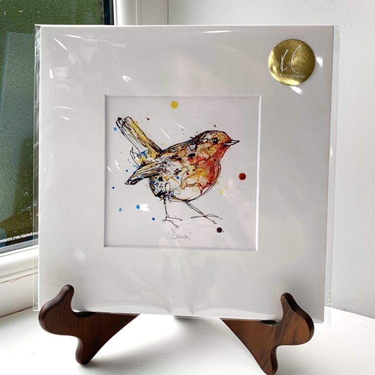 A miniature print of Dawn by Kathryn Callaghan, which features a bright eyed robin looking at the viewer. The print is presented mounted at 23cm, in a clear cello bag with a gold KC sticker in the top right corner, and is displayed here on a small wooden easel in a windowsill.