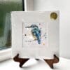 A miniature print of Daybreak by Kathryn Callaghan, which features a bright eyed kingfisher looking at the viewer. The print is presented mounted at 23cm, in a clear cello bag with a gold KC sticker in the top right corner, and is displayed here on a small wooden easel in a windowsill.