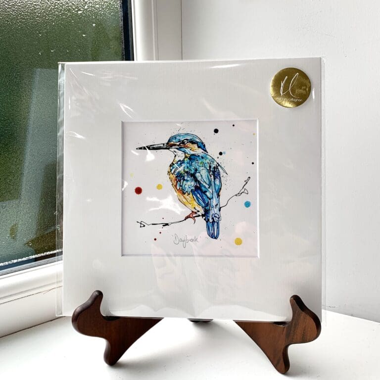 A miniature print of Daybreak by Kathryn Callaghan, which features a bright eyed kingfisher looking at the viewer. The print is presented mounted at 23cm, in a clear cello bag with a gold KC sticker in the top right corner, and is displayed here on a small wooden easel in a windowsill.