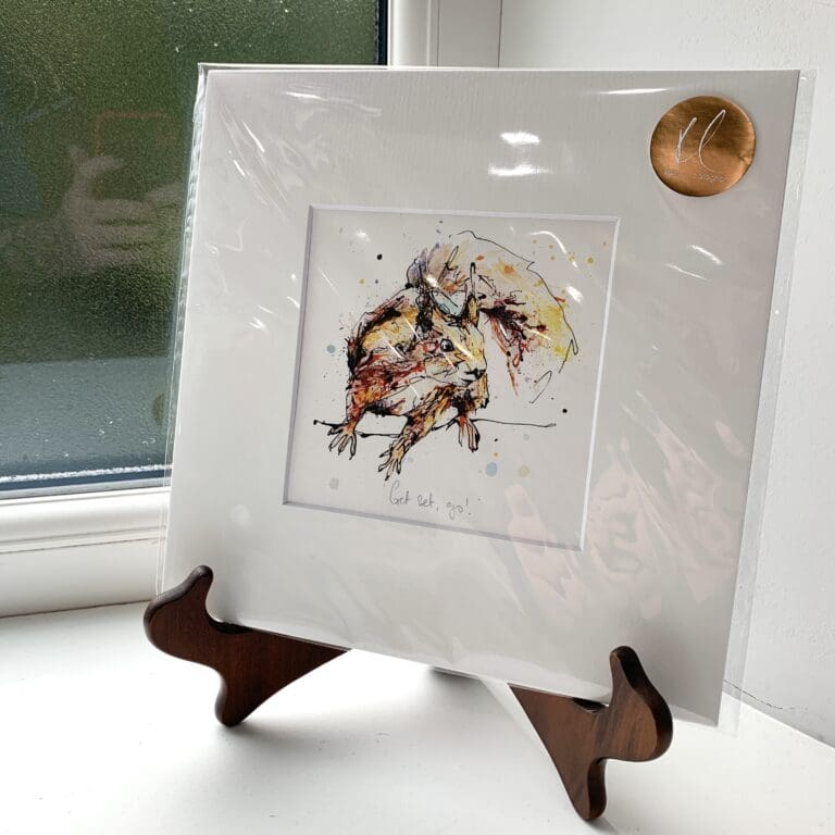 A miniature print of Get Set, Go! by Kathryn Callaghan, which features a red squirrel poised to run. The print is presented mounted at 23cm, in a clear cello bag with a gold KC sticker in the top right corner, and is displayed here on a small wooden easel in a windowsill.