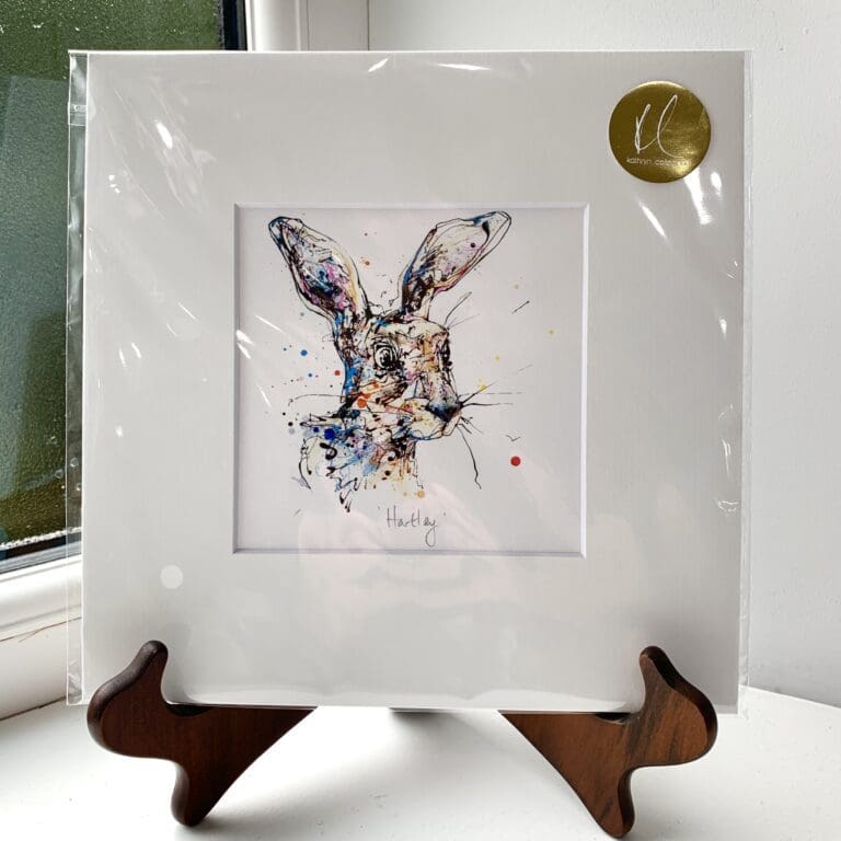 A miniature print of Hartley by Kathryn Callaghan, which features a frazzled looking hare's face. The print is presented mounted at 23cm, in a clear cello bag with a gold KC sticker in the top right corner, and is displayed here on a small wooden easel in a windowsill.