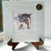 A miniature print of Hudson by Kathryn Callaghan, which features a friendly farmhorse looking over its fence. The print is presented mounted at 23cm, in a clear cello bag with a gold KC sticker in the top right corner, and is displayed here on a small wooden easel in a windowsill.