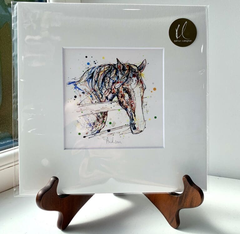 A miniature print of Hudson by Kathryn Callaghan, which features a friendly farmhorse looking over its fence. The print is presented mounted at 23cm, in a clear cello bag with a gold KC sticker in the top right corner, and is displayed here on a small wooden easel in a windowsill.