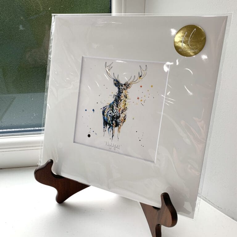 A miniature print of Nightfall by Kathryn Callaghan, which features a stately stag captured in dark blues and golds. The print is presented mounted at 23cm, in a clear cello bag with a gold KC sticker in the top right corner, and is displayed here on a small wooden easel in a windowsill.