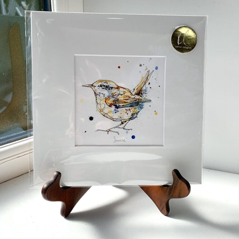 A miniature print of Sunrise by Kathryn Callaghan, which features a bright eyed wren poised charismatically with its tail held high. The print is presented mounted at 23cm, in a clear cello bag with a gold KC sticker in the top right corner, and is displayed here on a small wooden easel in a windowsill.