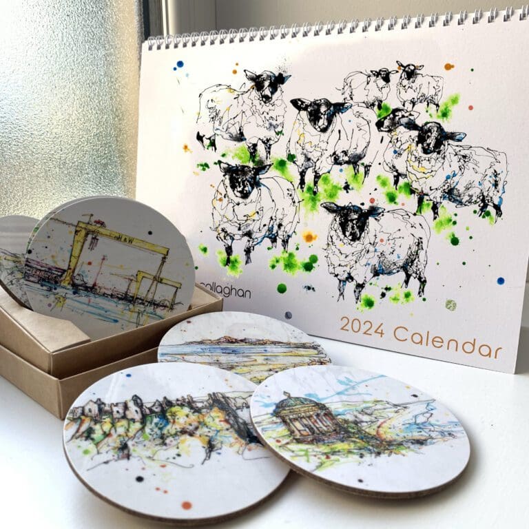 A 2024 calendar bearing a cover image of a painted flock of sheep, named Baa by Kathryn Callaghan, sits on a bright windowsill behind a set of scattered coasters, which bear artwork of landscapes around Northern Ireland, also by Kathryn Callaghan.