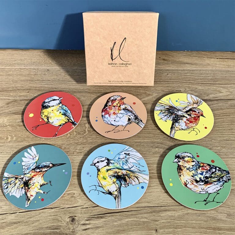 A set of six colourful coasters featuring garden bird prints by Kathryn Callaghan, including a red coaster with a blue tit, orange and yellow coasters featuring robins, a teal coaster featuring a kingfisher, a light blue coaster featuring a blue tit, and a green coaster featuring a chaffinch.