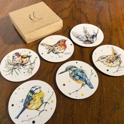 A set of six coasters featuring prints of small birds by Kathryn Callaghan, alongside a branded card box, sitting on a dark wood table. The birds featured are a goldfinch, robin, swallow, blue tit, kingfisher, and wren.