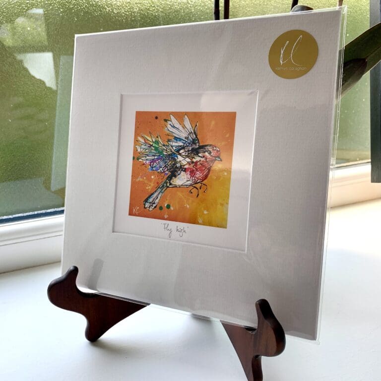 A miniature print of Fly High by Kathryn Callaghan, which features a robin in flight against a bright orange and yellow background, surrounded by a thin border of white where the print's title is printed from a pencil handwritten title. The print is presented mounted at 23cm, in a clear cello bag with a gold KC sticker in the top right corner, and is displayed here on a small wooden easel in a windowsill.