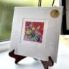 A miniature print of Love by Kathryn Callaghan, which features a posie of flowers on a vivid red background, surrounded by a thin border of white where the print's title is printed from a pencil handwritten title. The print is presented mounted at 23cm, in a clear cello bag with a gold KC sticker in the top right corner, and is displayed here on a small wooden easel in a windowsill.
