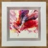 An original abstract painting by Kathryn Callaghan, named Passion. The painting features a swell of red, purple, yellow, and pink paint blown from the lower third of the surface board, and is framed in a deep floating white wood frame.