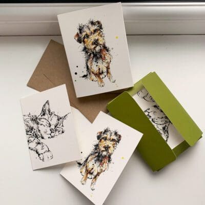 A set of miniature notecards featuring prints by Kathryn Callaghan. The set is opened to show the viewer the paper sleeve featuring Expectation, a terrier print, the green paper wallet that holds the cards inside, both card designs - Expectation and a print of a sleeping cat named Dreamer - and their accompanying cards.