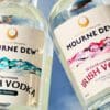 Two bottles of Mourne Dew Distillery Irish vodka photographed against mirrored surface reflecting a cloudy blue sky. Both labels feature the same artwork of Kathryn Callaghan's Essence of the Mournes print. The left bottle's label is blue and bears the text "Essence of Mourne Mourne Dew Premium Irish Vodka 6x Distilled". The right is pink and reads "Essence of Mourne Mourne Dew Wild Mourne Rose Irish Vodka 6x Distilled with the finest Irish rose petals".
