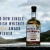 A promotional image of Mourne Dew Distillery's Single Malt Irish Whiskey, featuring the bottle sat on the wall of Spelga Dam in Northern Ireland. The bottle's label features Kathryn Callaghan's Essence of the Mournes print, and digitally added text beside the bottle reads "Mourne Dew Single Malt Irish Whiskey IWSC Gold Award Winner." The IWSC Gold Label Graphic also features in the top right.