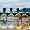 Four bottles of Mourne Dew Distillery spirits laid on a wooden cheeseboard in a field in the Mourne Mountains. From left to right the bottles are Kilbroney Gin, Vodka, Whiskey, and Poitín. The first three feature Kathryn Callaghan's Essence of the Mournes print, and the Poitín features her Púca print.