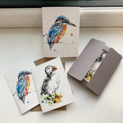 A set of miniature notecards featuring prints by Kathryn Callaghan. The set is opened to show the viewer the paper sleeve featuring Gaze, a kingfisher print, the grey paper wallet that holds the cards inside, both card designs - Gaze and another print named Atlantic puffin - and their accompanying cards.