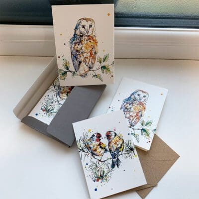 A set of miniature notecards featuring prints by Kathryn Callaghan. The set is opened to show the viewer the paper sleeve featuring Curio, a barn owl print, the green paper wallet that holds the cards inside, both card designs - Curio and a print of a goldfinch pair named I Turn To You - and their accompanying cards.