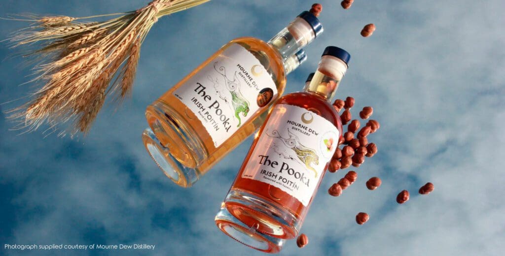 Two bottles of Mourne Dew Distillery Irish Poitín photographed against a mirrored surface reflection a cloudy blue sky. The surface is littered with hazelnuts and wheat, and the label bears Kathryn Callaghan's Púca painting.