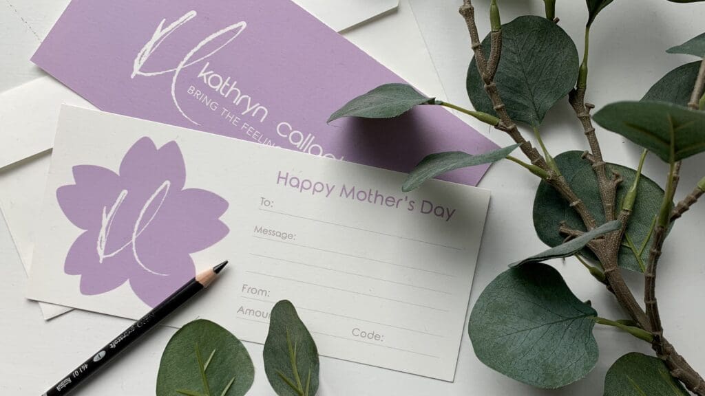 A promotional image of Kathryn Callaghan's Mother's Day Gift Voucher. The vouchers have a lilac back and Kathryn's logo and company name, and on the front have a lilac blossom shape bearing the logo and space for a dedication name and message, price amount, and code. The voucher in this photo is surrounded by eucalyptus leaves and a pencil.