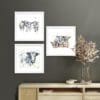 Three prints by Kathryn Callaghan shown in white frames in situ above a pine sideboard. The prints are A Good Natter, which shows three dairy cows standing together, Pastures New, a single dairy cow standing looking at the viewer, and Well, Hello, which shows three dairy cows looking over a red fence, shown in Standard White Frames in Situ.