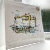 A print of Belfast Docks by Kathryn Callaghan, which features yellow Harland & Wolff cranes set before Cave Hill. The 30cm print is presented flat in a clear cello bag with a golden KC sticker in the top right.